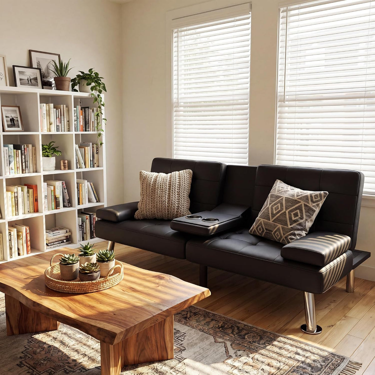 Staged living room with a black futon sofa bed, center cup-holder arm, pillows, bookcase and wooden coffee table.