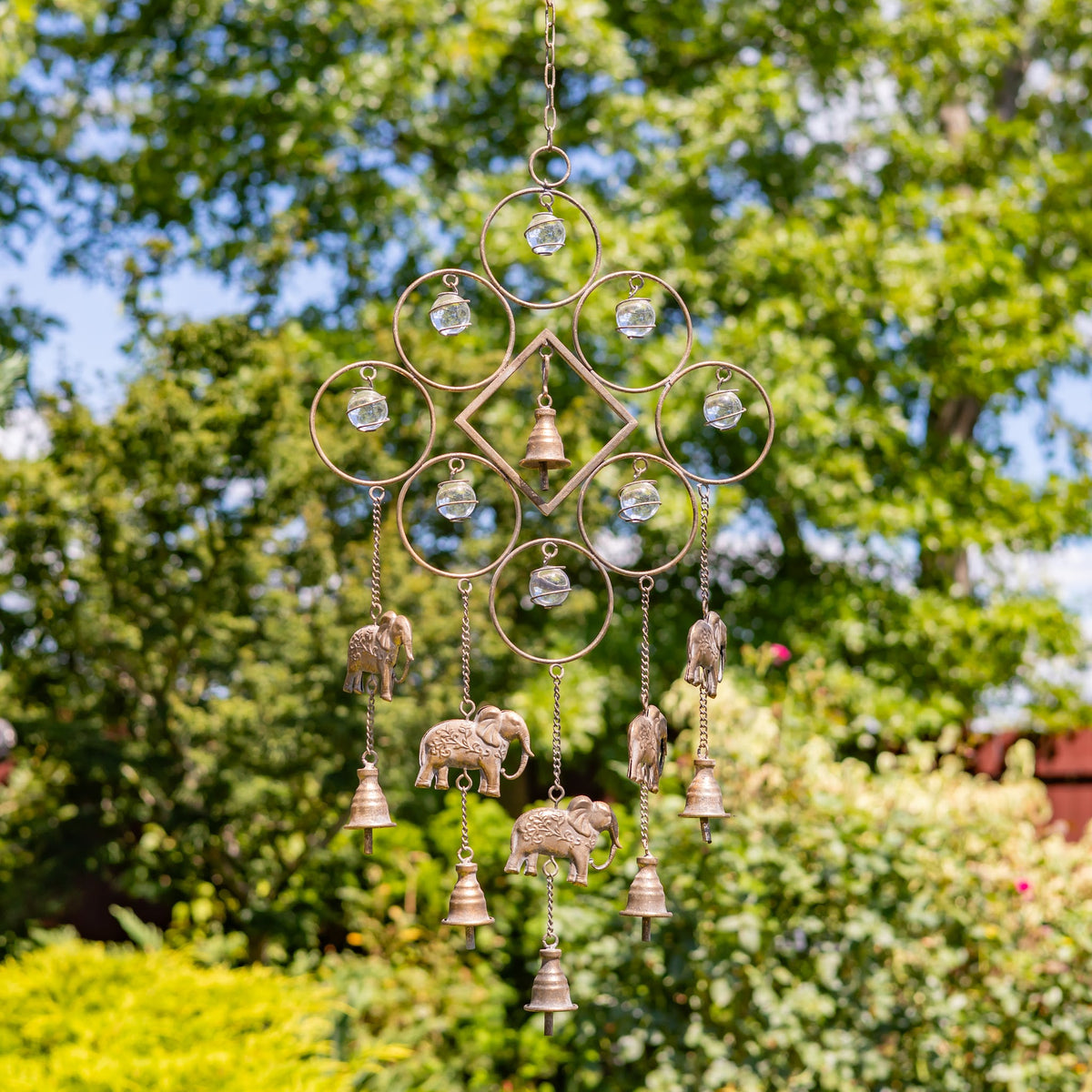 Metal wind chime with circular wirework, dangling bells and elephant ornaments hanging outdoors among green trees.