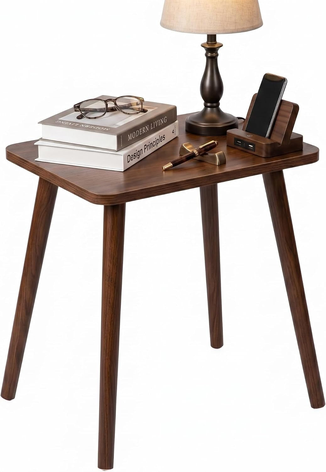 Small rectangular wood side table with tapered legs holding a lamp, stacked books, glasses, pen, and a phone dock