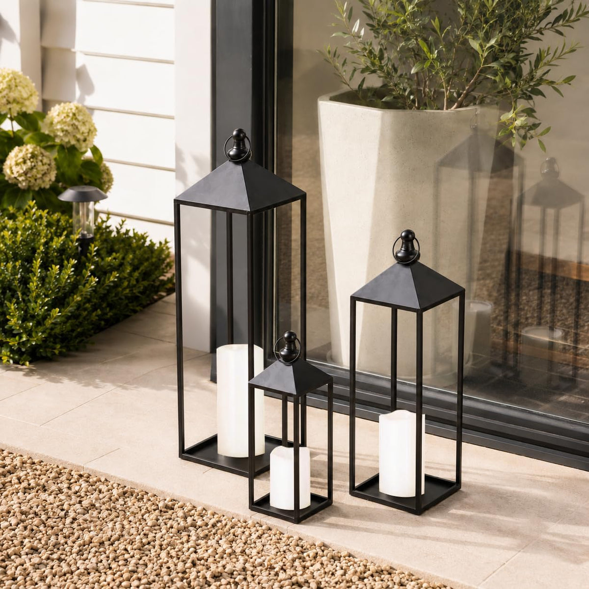 Three black metal lanterns with LED candles placed on a patio floor by a sliding glass door in daylight