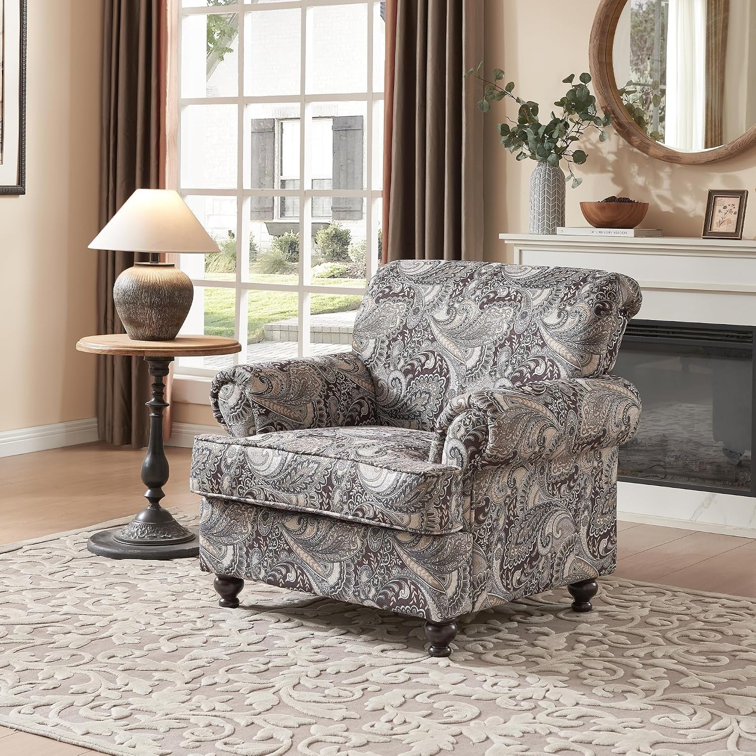 Side view of a paisley-patterned upholstered armchair beside a round wooden side table and lamp.