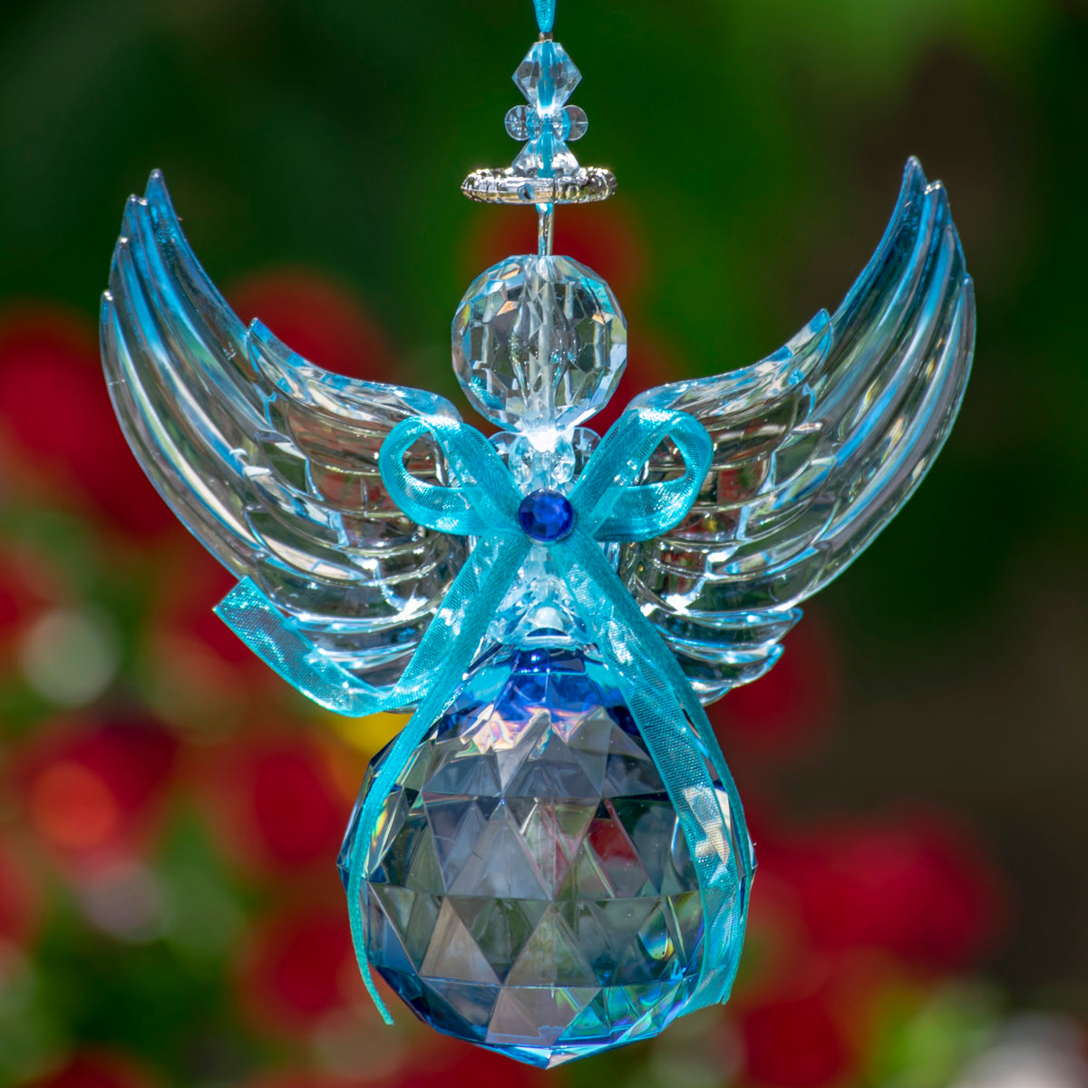 A blue and clear faceted angel ornament hangs against a blurred green and red background.