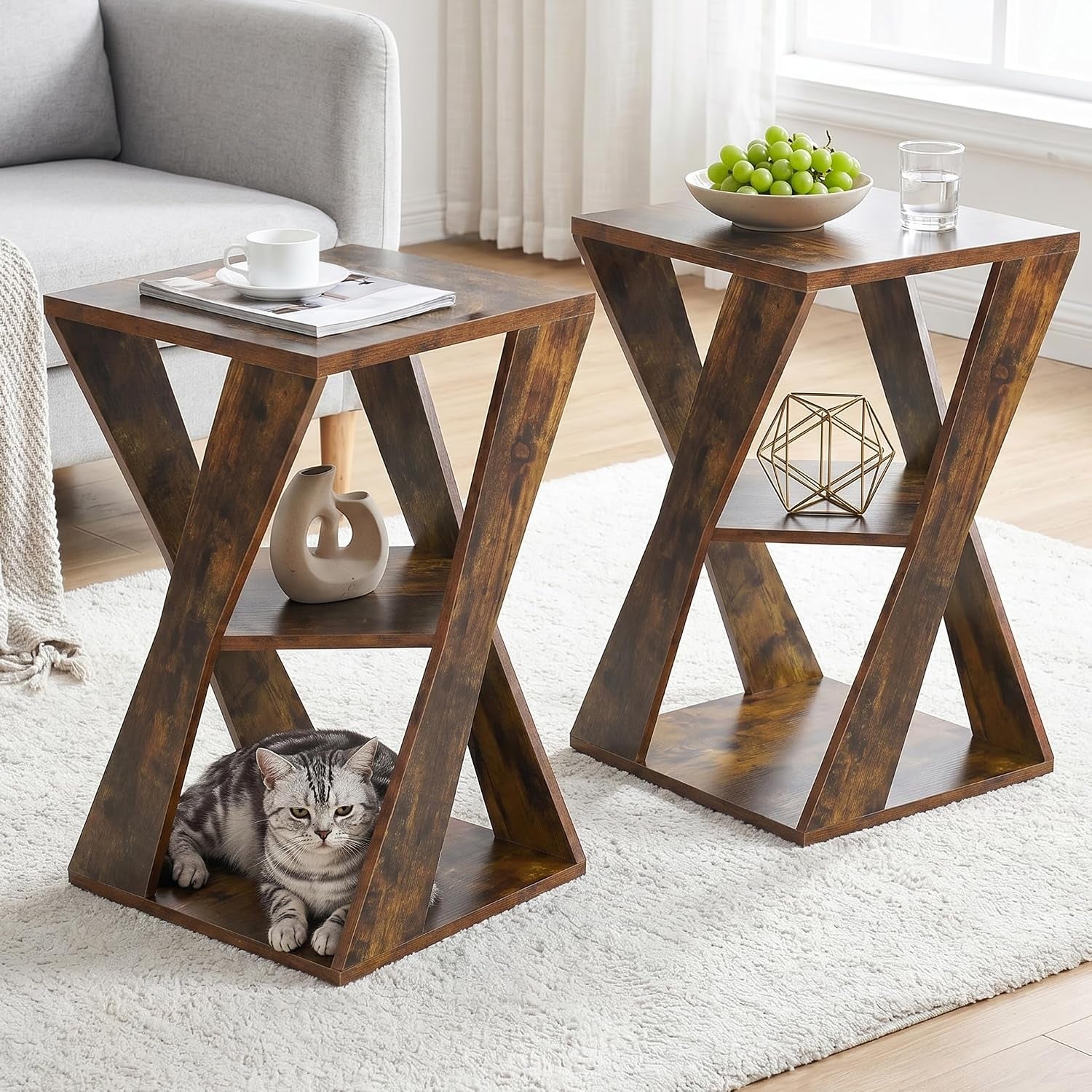 Two matching X-shaped wooden side tables staged on a white rug next to a sofa, one with a cat underneath.