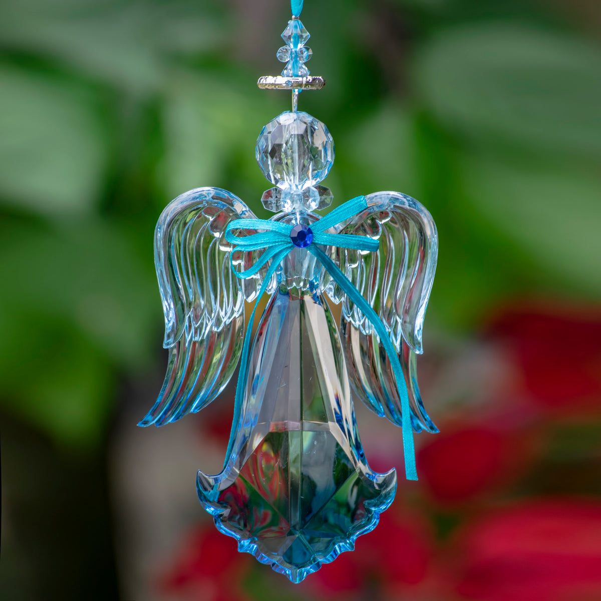 Clear faceted hanging angel with curved wings, a turquoise ribbon bow and blue-edged dress against blurred greenery.