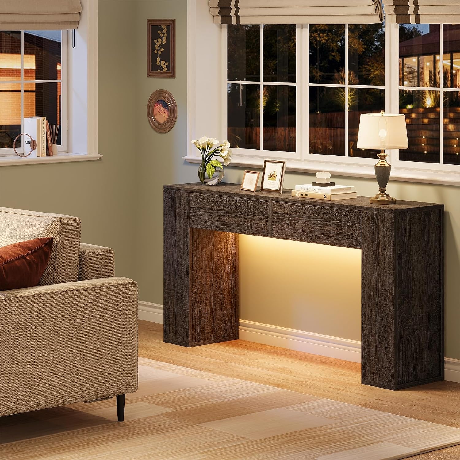 Angled living-room view of a grey console table under a window with a lamp, framed photos and warm LED glow.