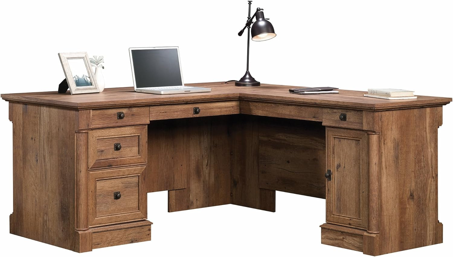L-shaped wooden desk in a warm brown finish staged with a laptop and lamp, showing three left drawers and a right cabinet.