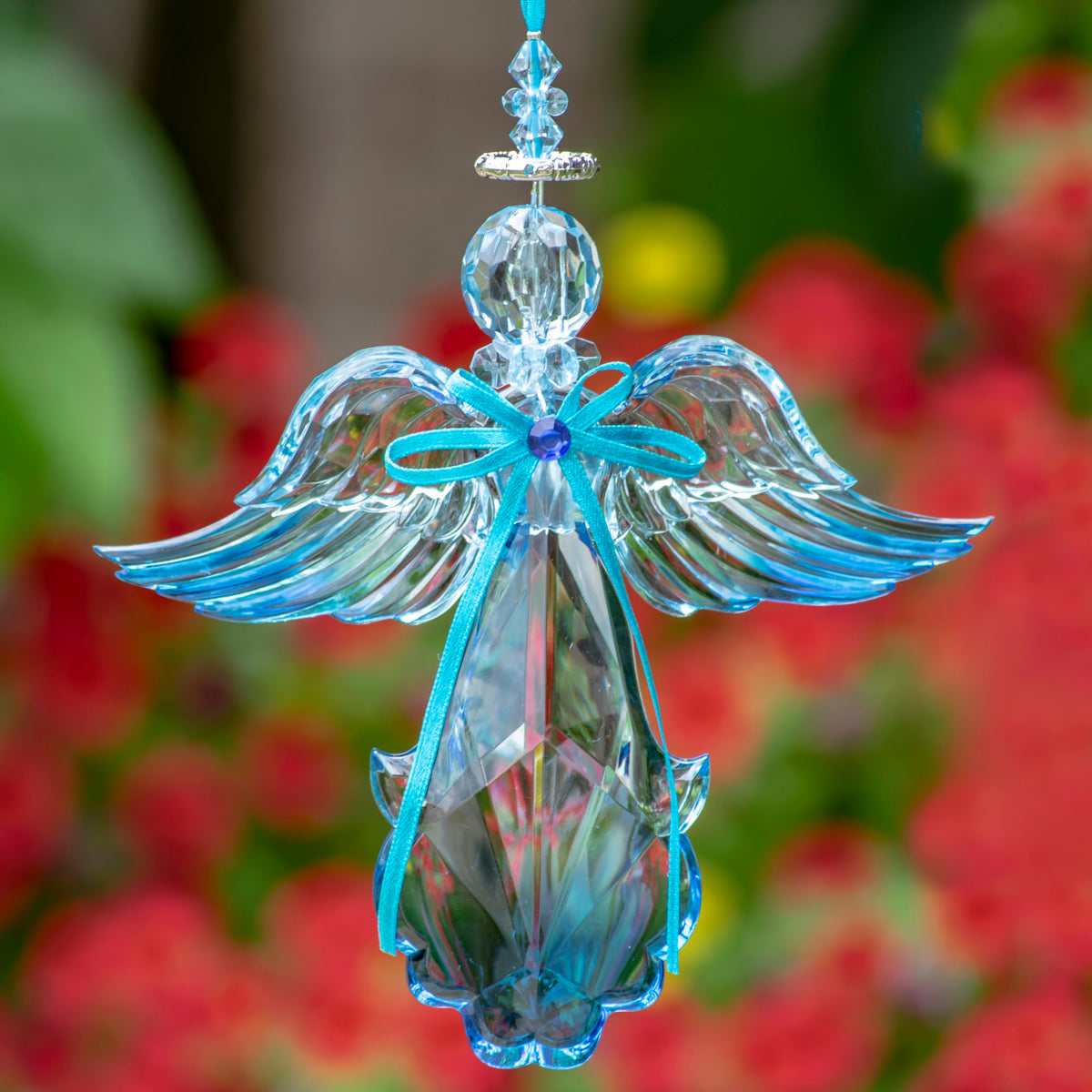 Blue faceted angel ornament with outstretched wings and a turquoise ribbon, hanging against blurred flowers.