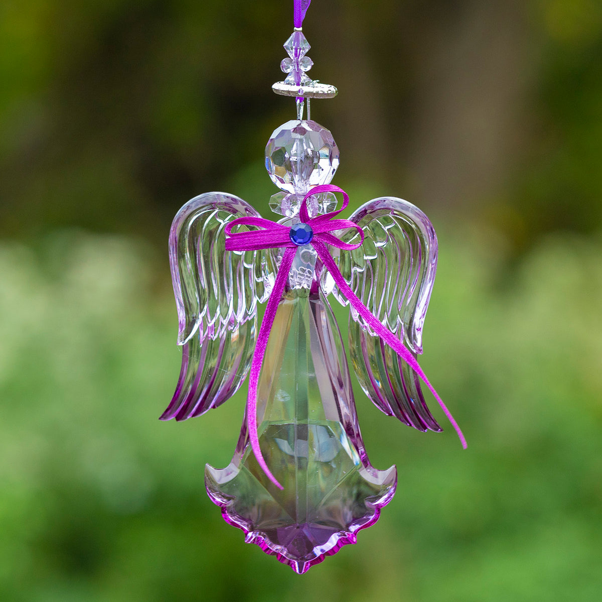 Purple-toned faceted angel ornament with curved wings and a bright ribbon bow, hanging against a blurred green background.