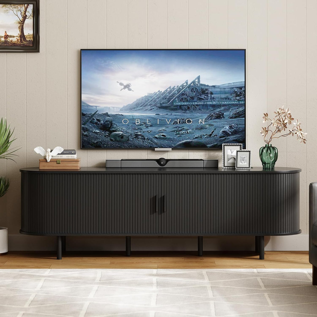 Wide living-room composition with the black TV stand under a large wall TV, plant and framed photos beside it.