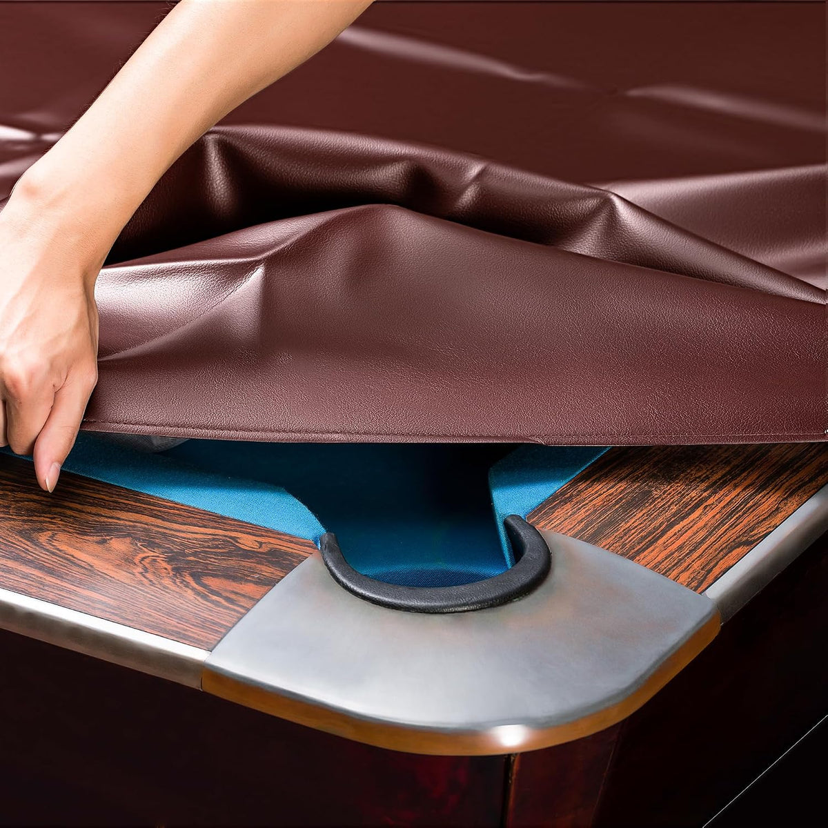 Hand lifting a brown leatherette cover from a pool table corner, revealing blue felt pocket and wood trim