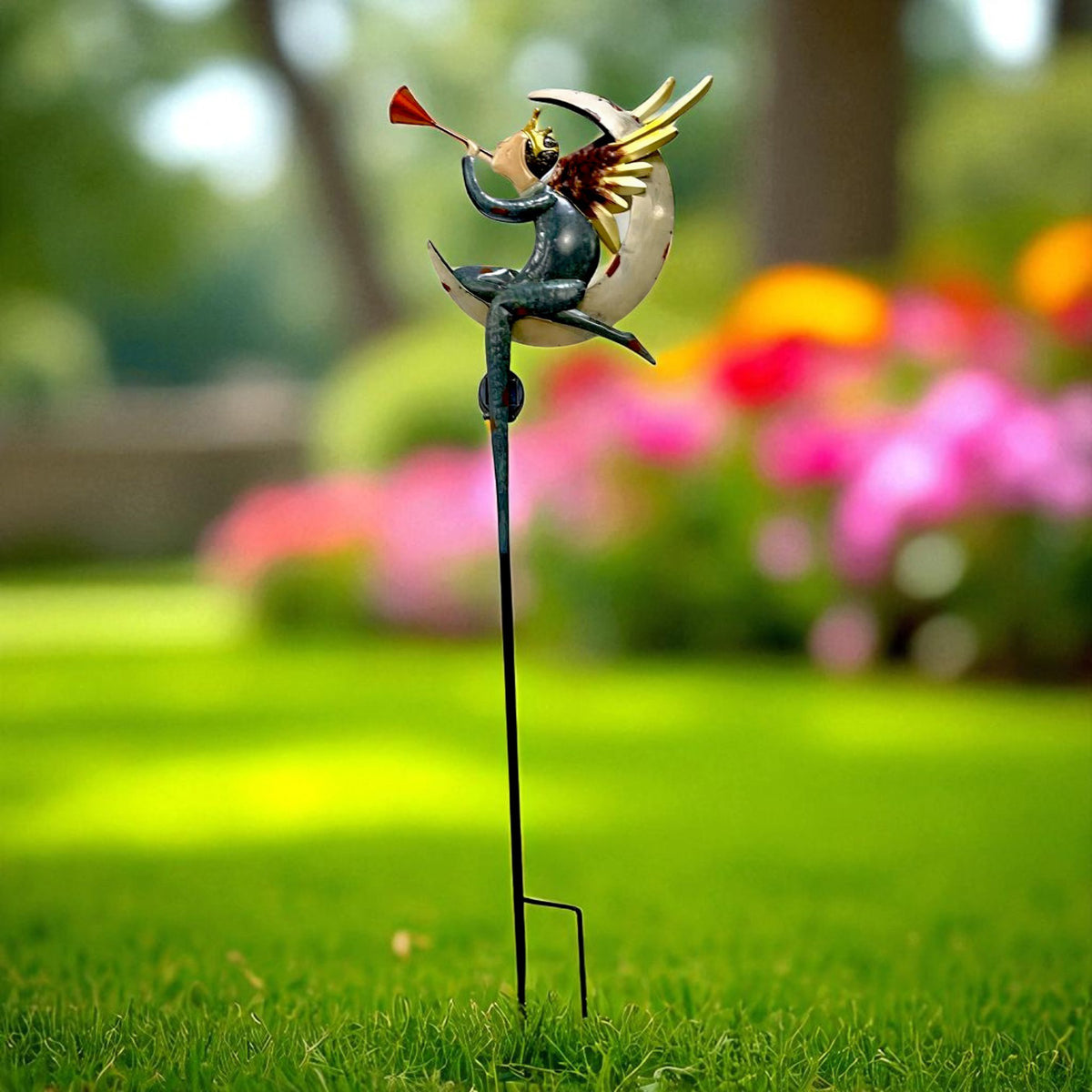 A blue-toned angel stake blows a small trumpet above a lawn, with flowers blurred in the background.