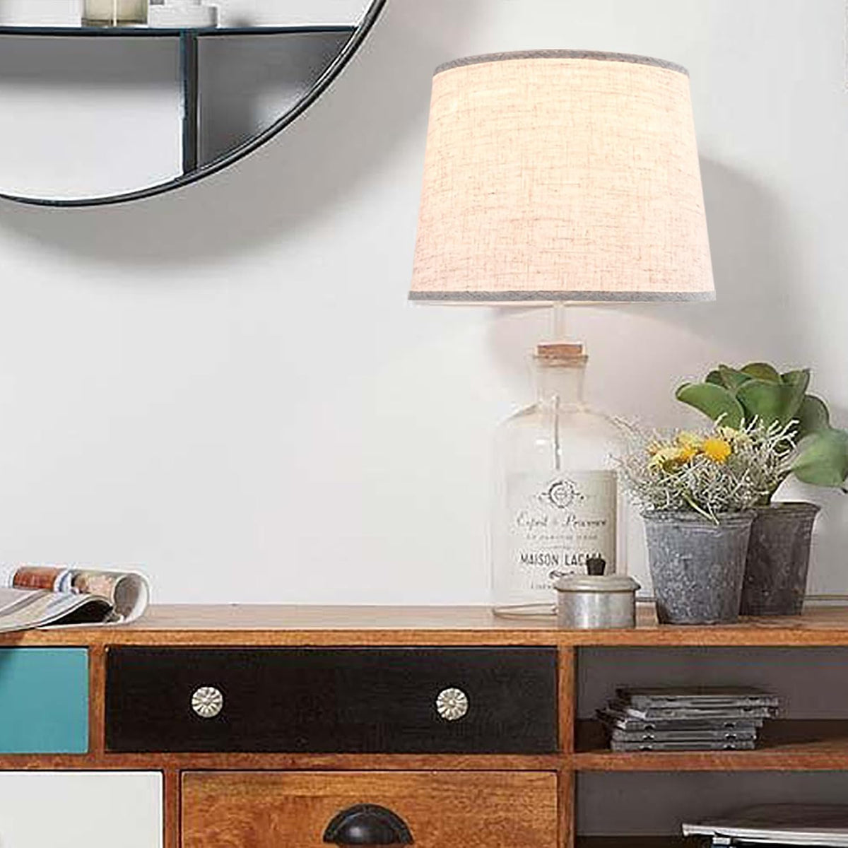 Table lamp with a warm oatmeal linen drum shade on a wood sideboard with plants and mirror.