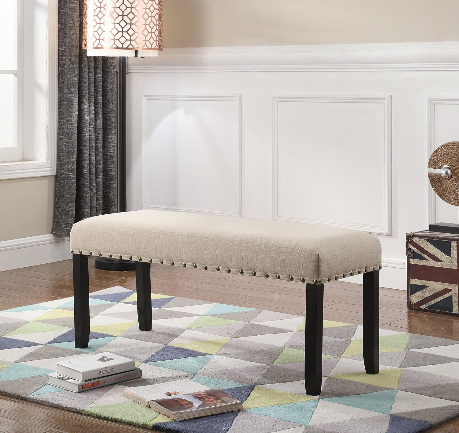 Tan upholstered dining bench in a room setting on a geometric rug, side view showing nailhead trim and dark legs