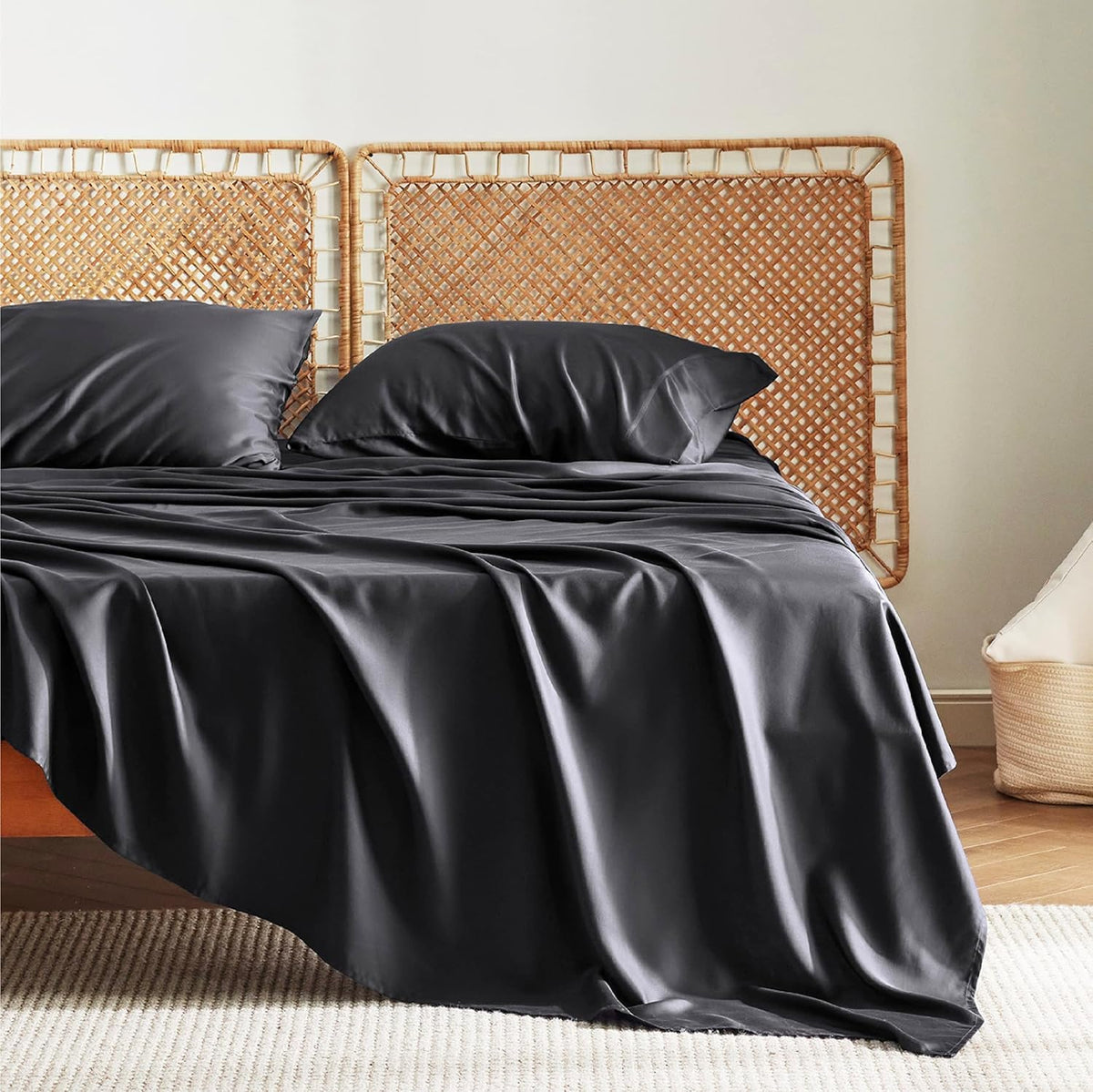 Three-quarter view of a bed with dark charcoal silky sheets and two pillows against a woven cane headboard.