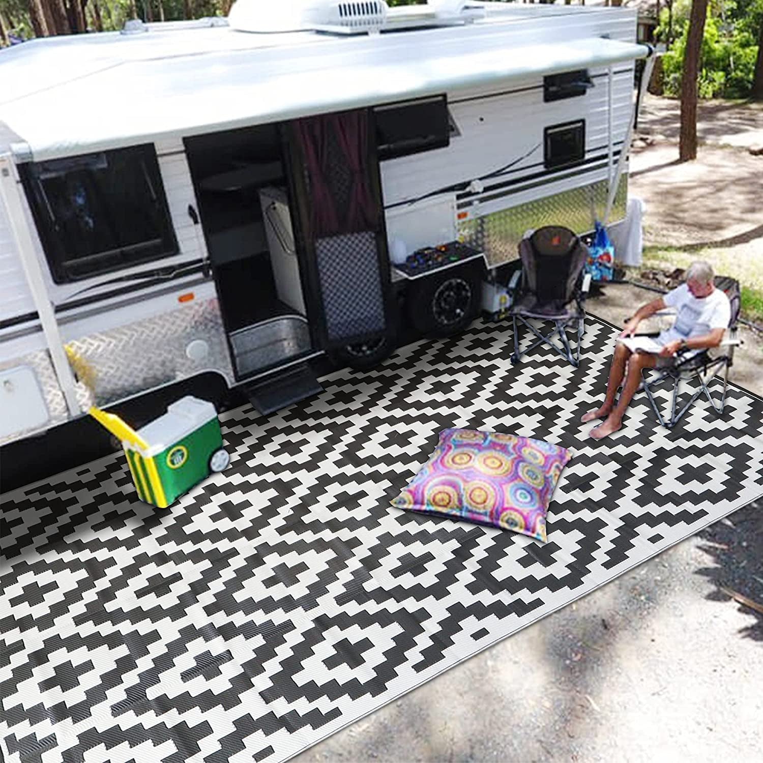 Camp setup outside an RV with a large black-and-white patterned mat, folding chairs and cooler.