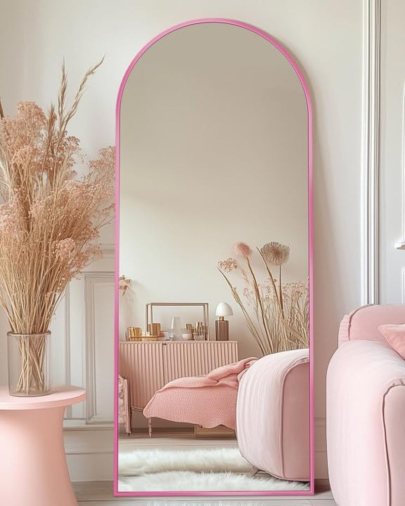 Tall arched full-length mirror with a pink frame leaning against a white wall next to pink furniture.
