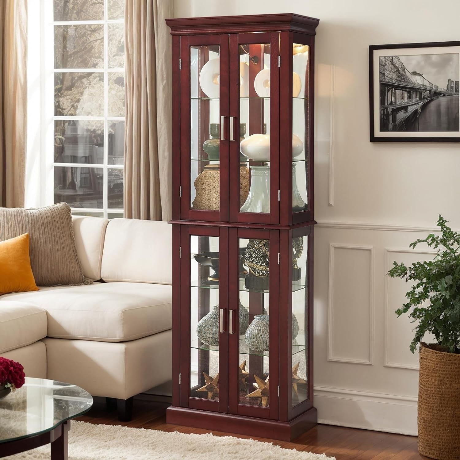 Tall dark cherry curio cabinet with glass doors and interior lighting placed in a living room corner beside a light sofa.