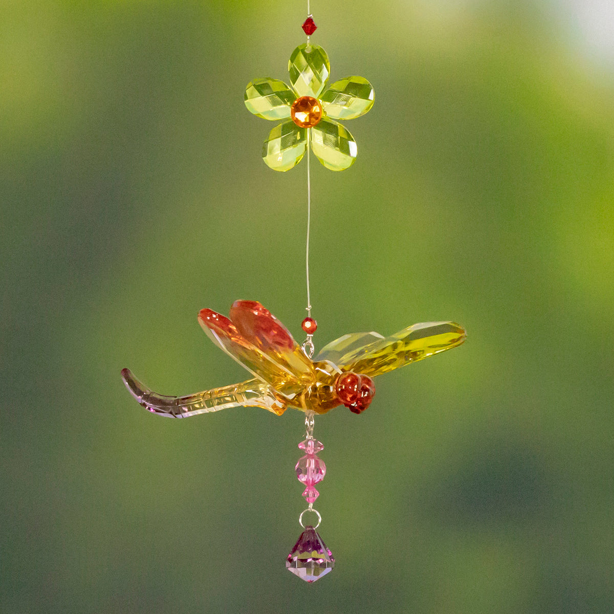 A colorful faceted dragonfly ornament hangs below a green flower against a blurred green background.