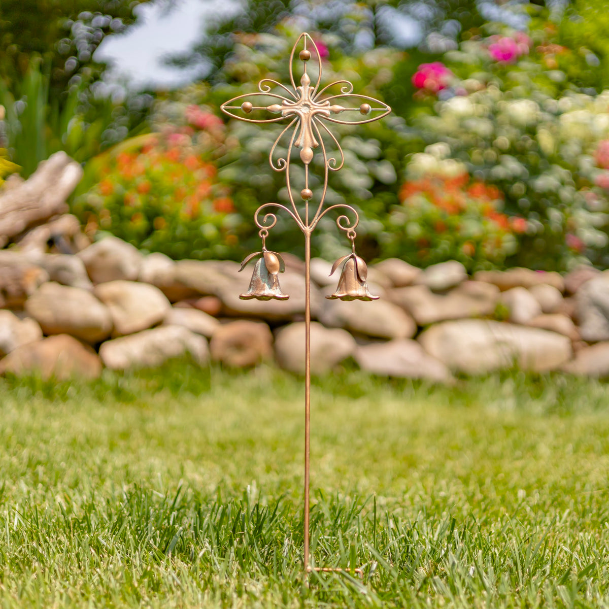 Ornate metal cross garden stake with two dangling bell-shaped ornaments in a grassy garden.