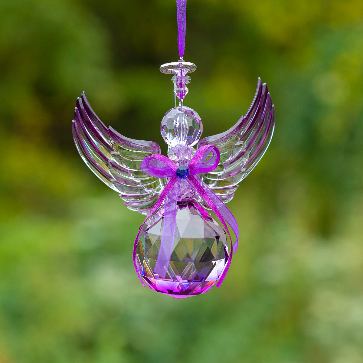 A faceted purple acrylic angel ornament with raised wings and a ribbon bow hangs against a blurred green background.