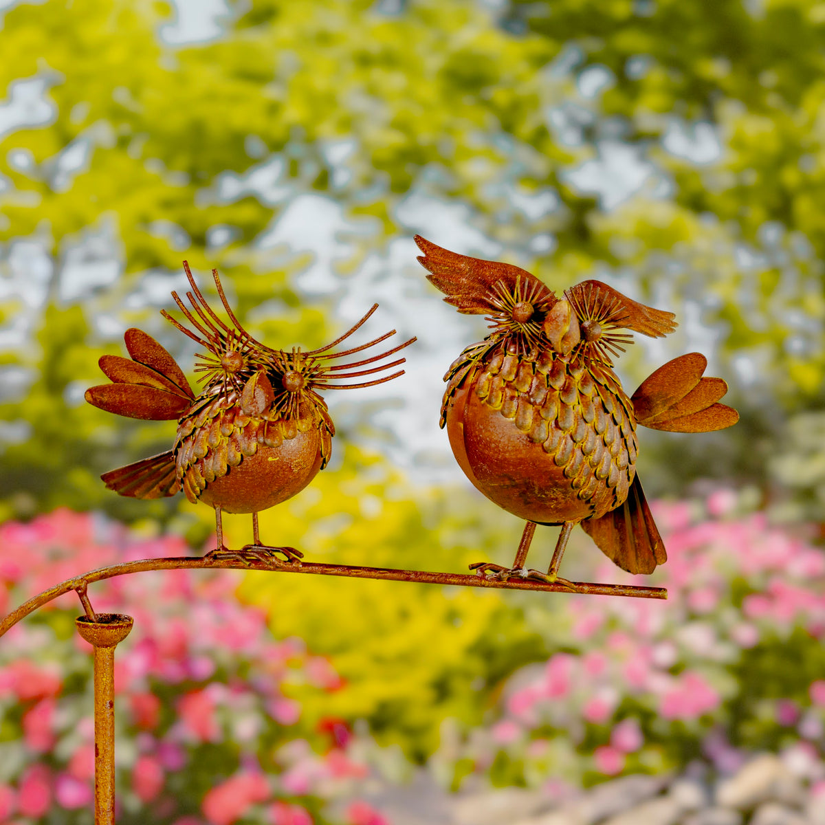 Two rust-colored metal birds balance on a curved garden stake against blurred flowers and foliage.