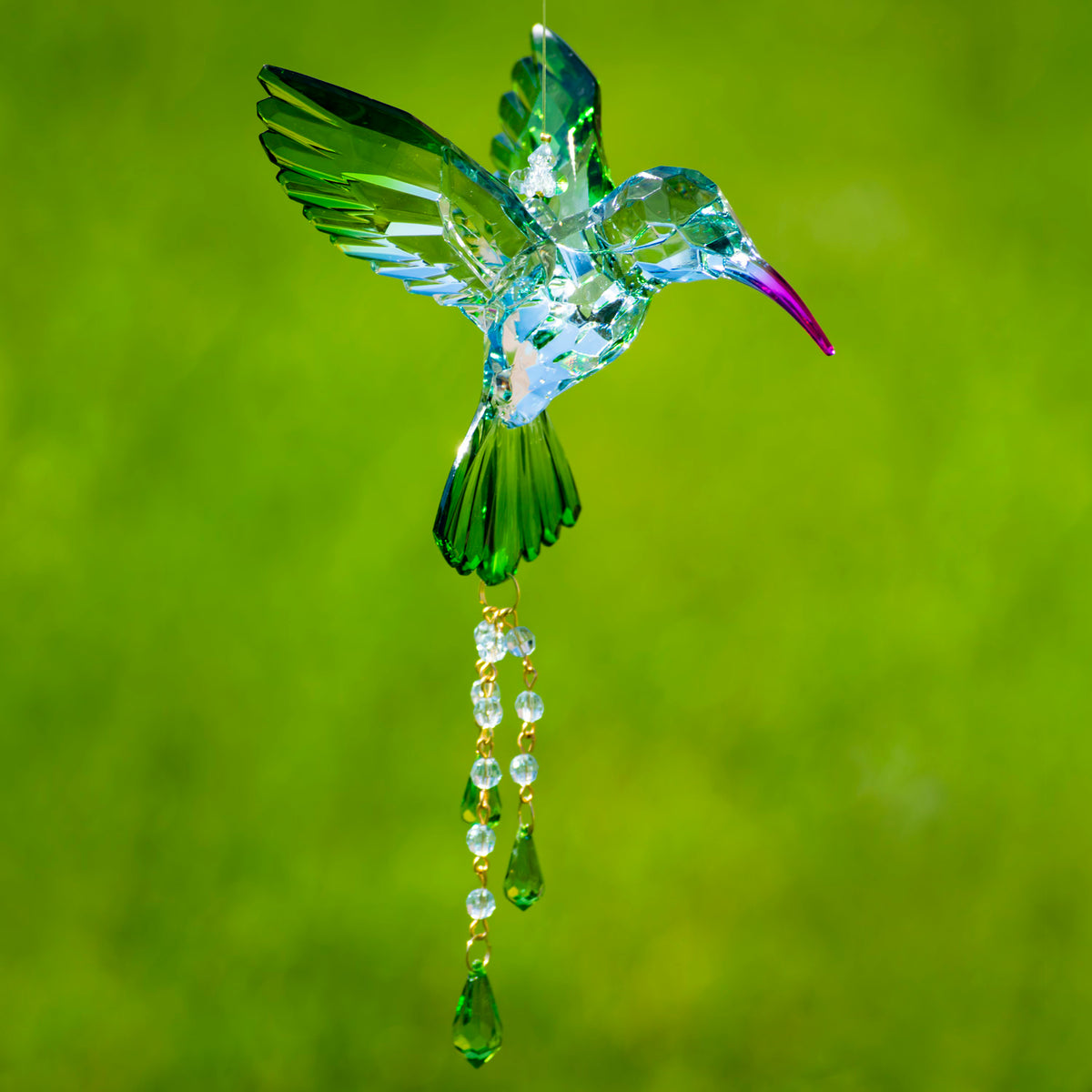 Green and blue faceted hummingbird ornament with dangling bead strands against a blurred green background.