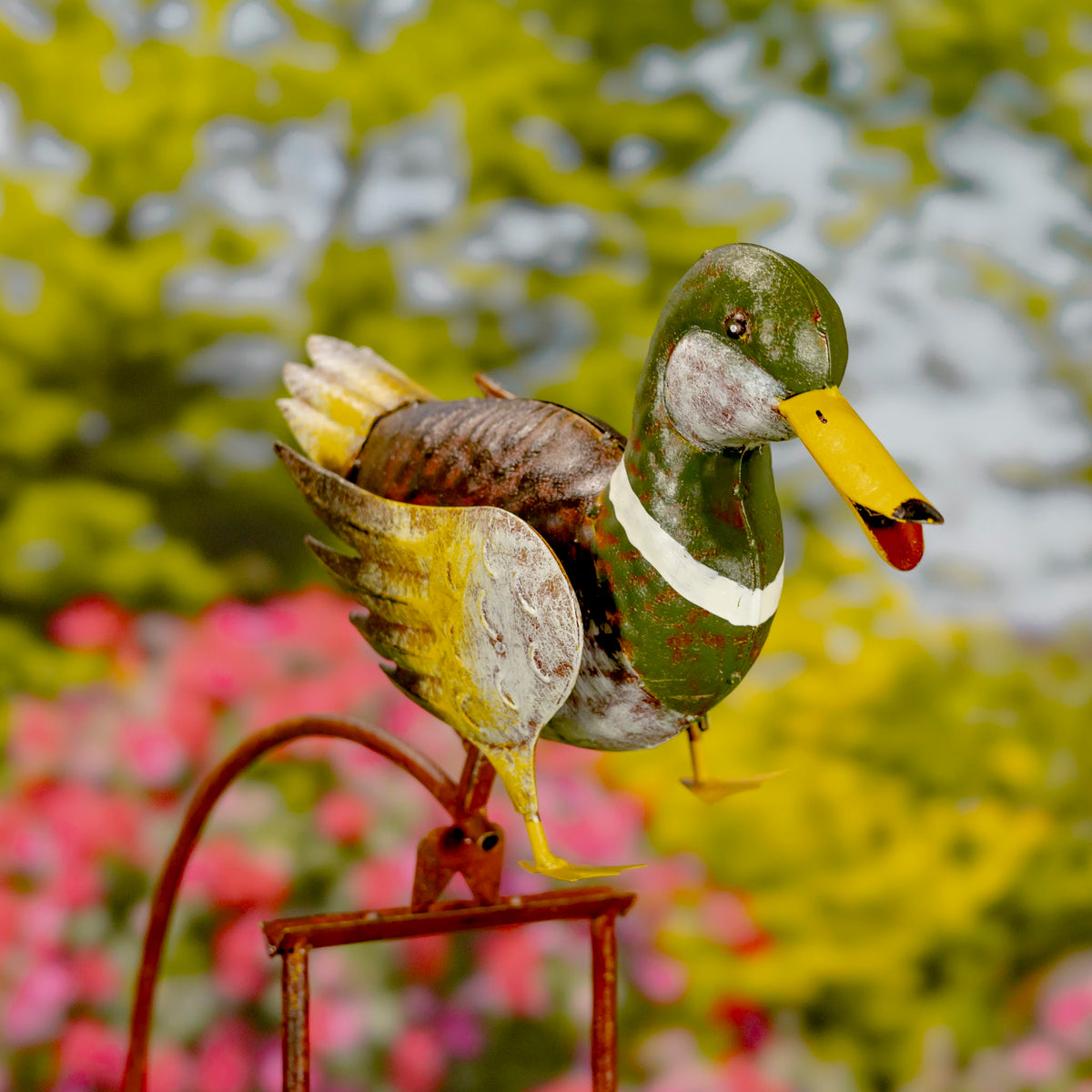 Painted mallard duck with outstretched wings perched on a curved metal rocking stake among flowers.