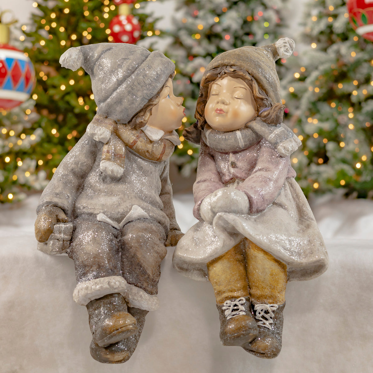 Two glittery winter-clad figurines sit side by side before a decorated Christmas tree.
