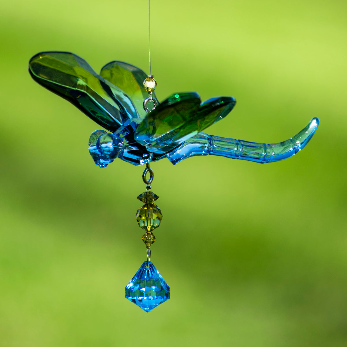 Blue and green acrylic dragonfly ornament with dangling dark beads and a blue faceted drop against a blurred green