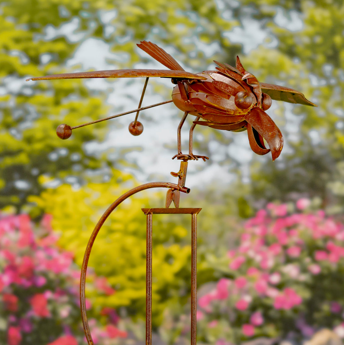 Rust-colored metal hawk stake with outstretched wings and hanging rods, shown against a blurred garden.