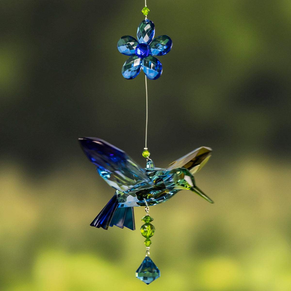 A blue-green faceted hummingbird ornament hangs below a blue flower, with a blue drop bead beneath.