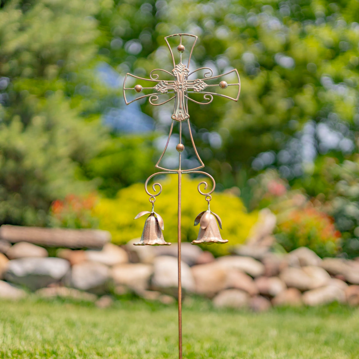 Ornate metal cross garden stake with two hanging bells standing in grass beside a rock border.