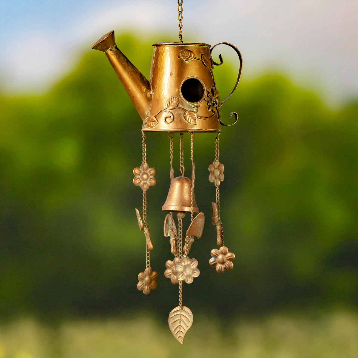 Antique-gold watering-can birdhouse wind chime hanging outdoors, with dangling flower and leaf ornaments.