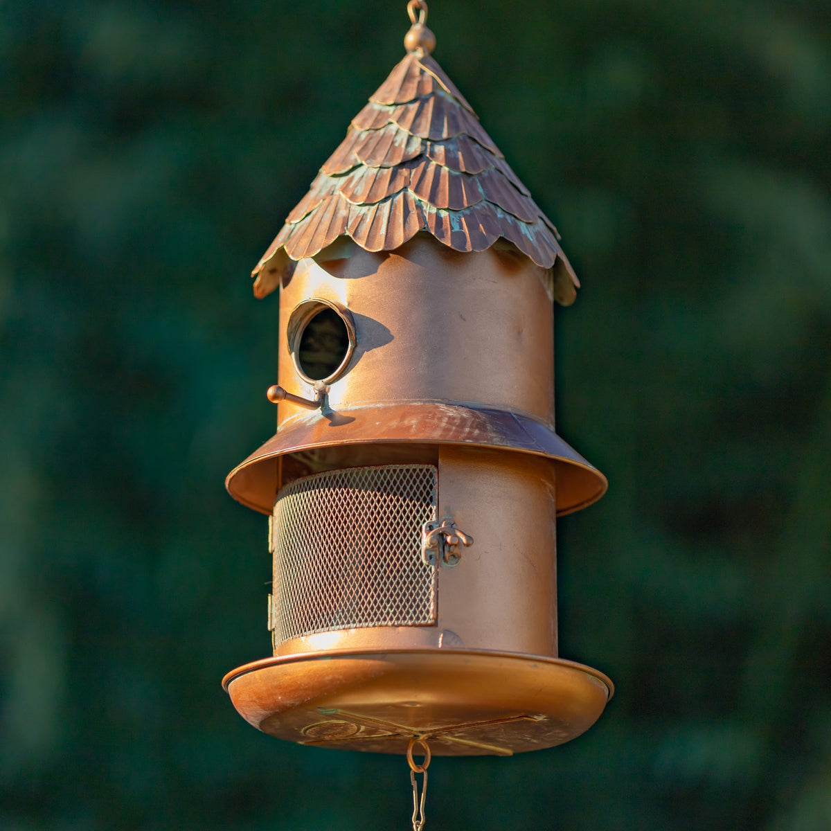 Copper-toned metal birdhouse hangs against a dark green background, with a mesh feeder and latched door.