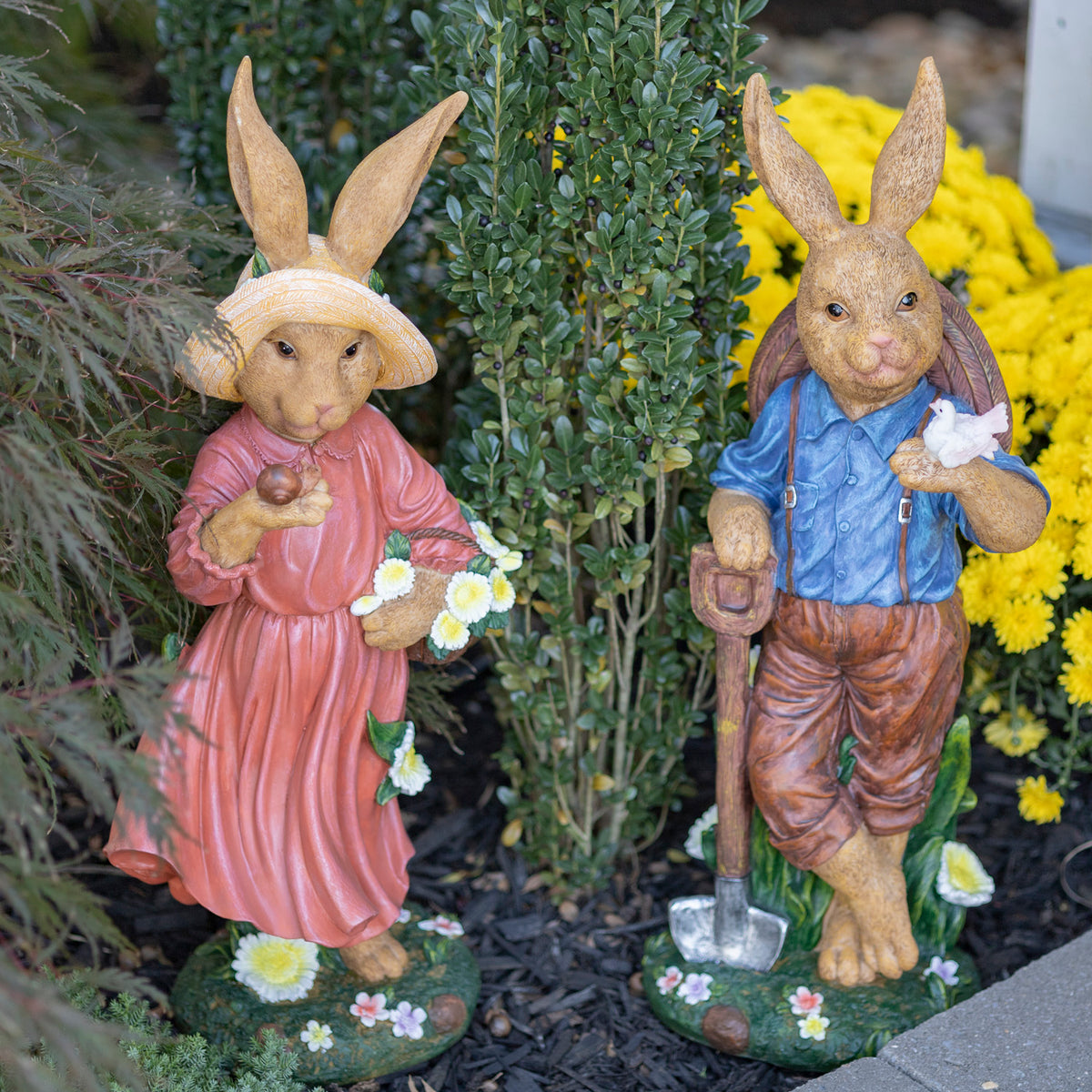 Rabbit statues in a garden: one in a blue shirt holding a bird and shovel, beside one in a pink dress.