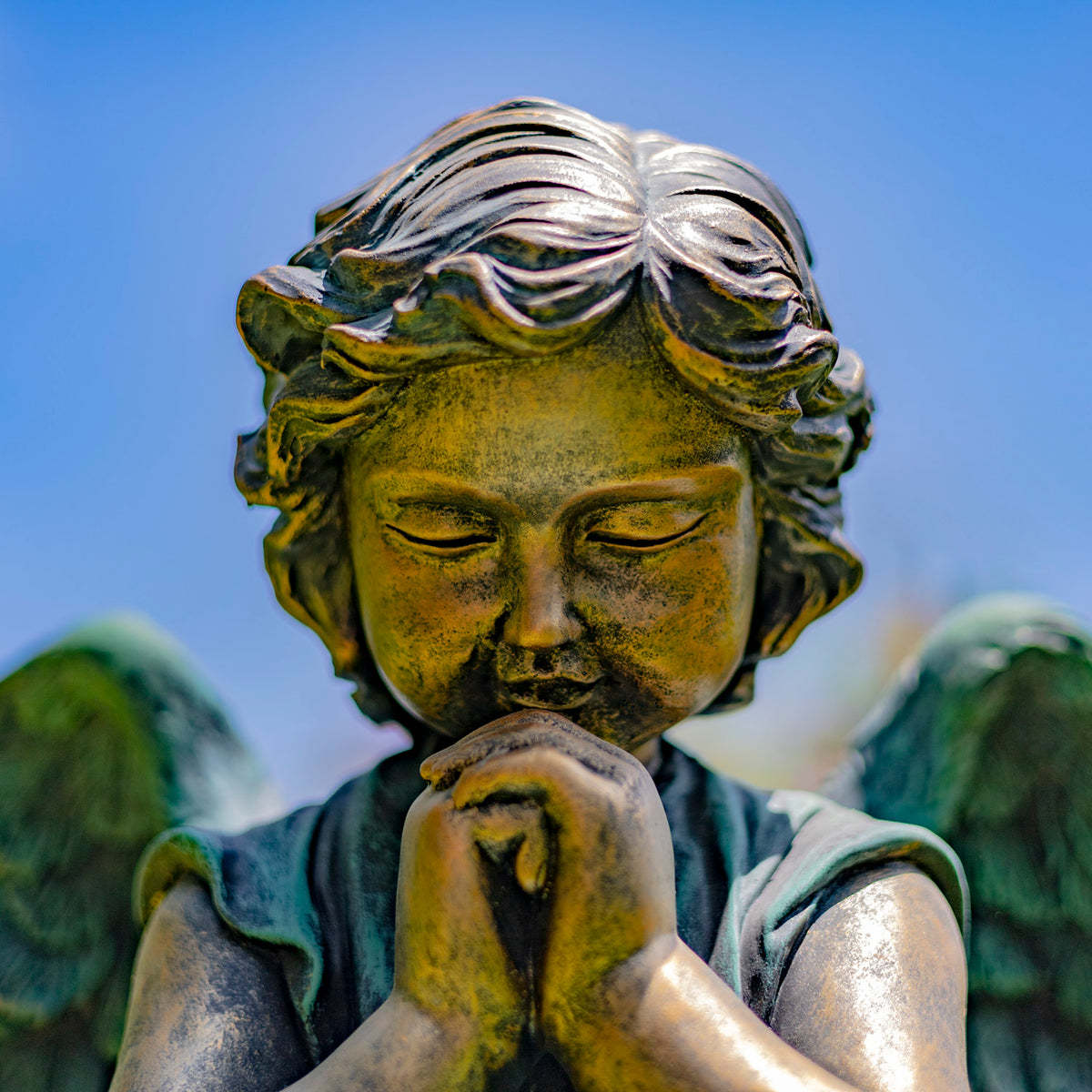 Close-up of the angel’s bowed face, clasped hands, and upper wings against a blue sky.