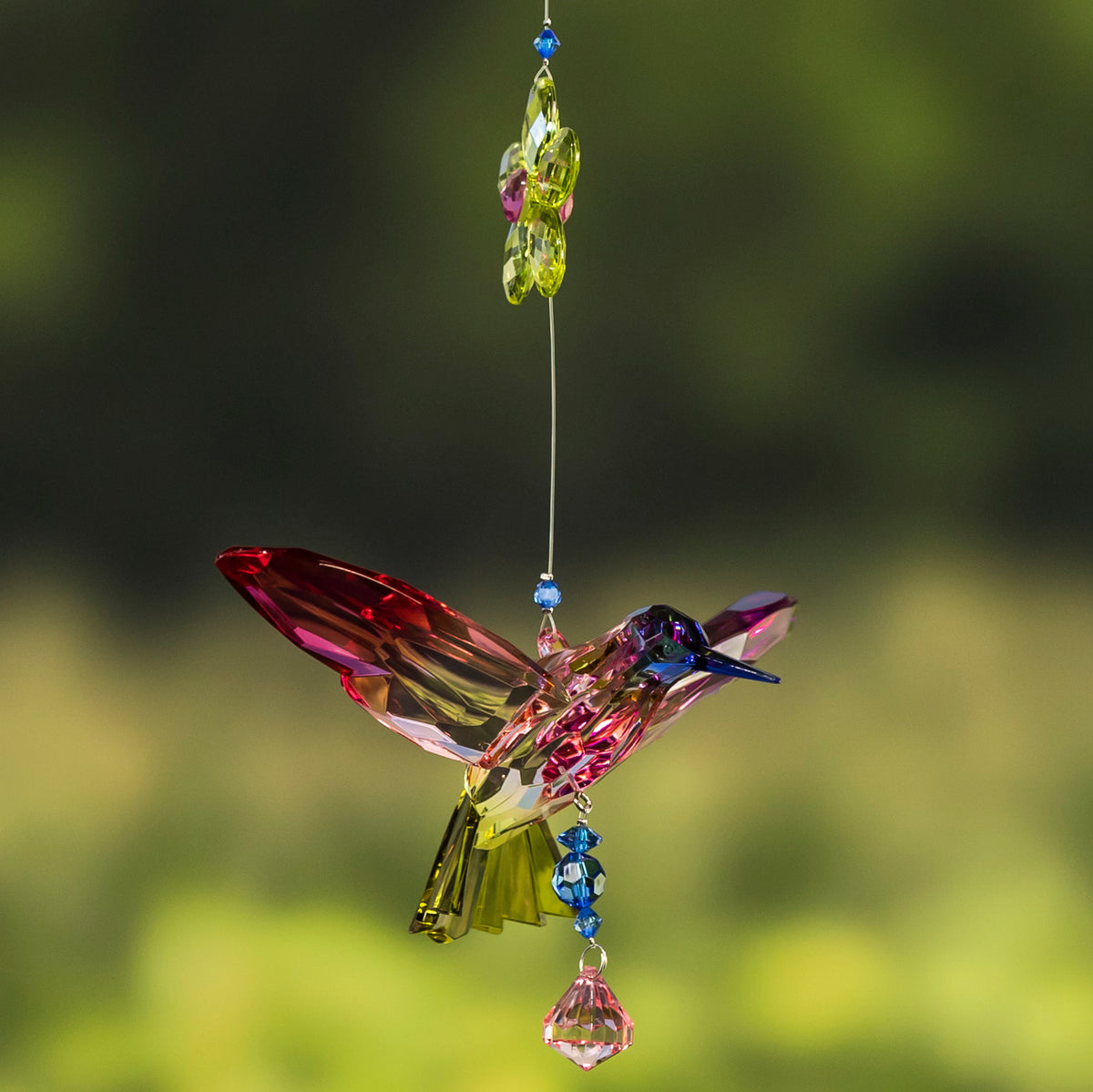 Pink and green faceted hummingbird ornament hangs from a beaded strand against a blurred green background.