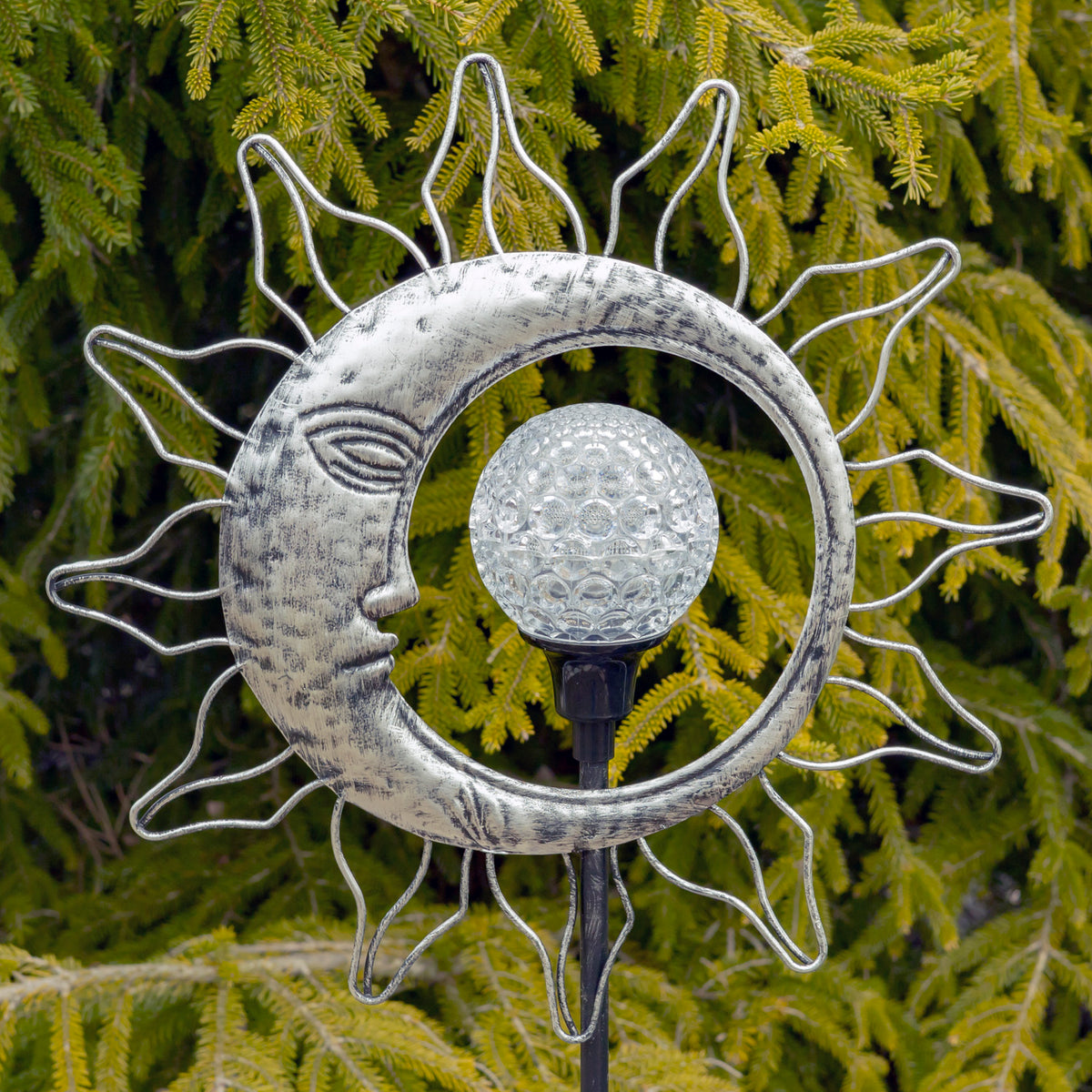 Silver sun-and-moon garden stake with a clear globe among evergreen branches.