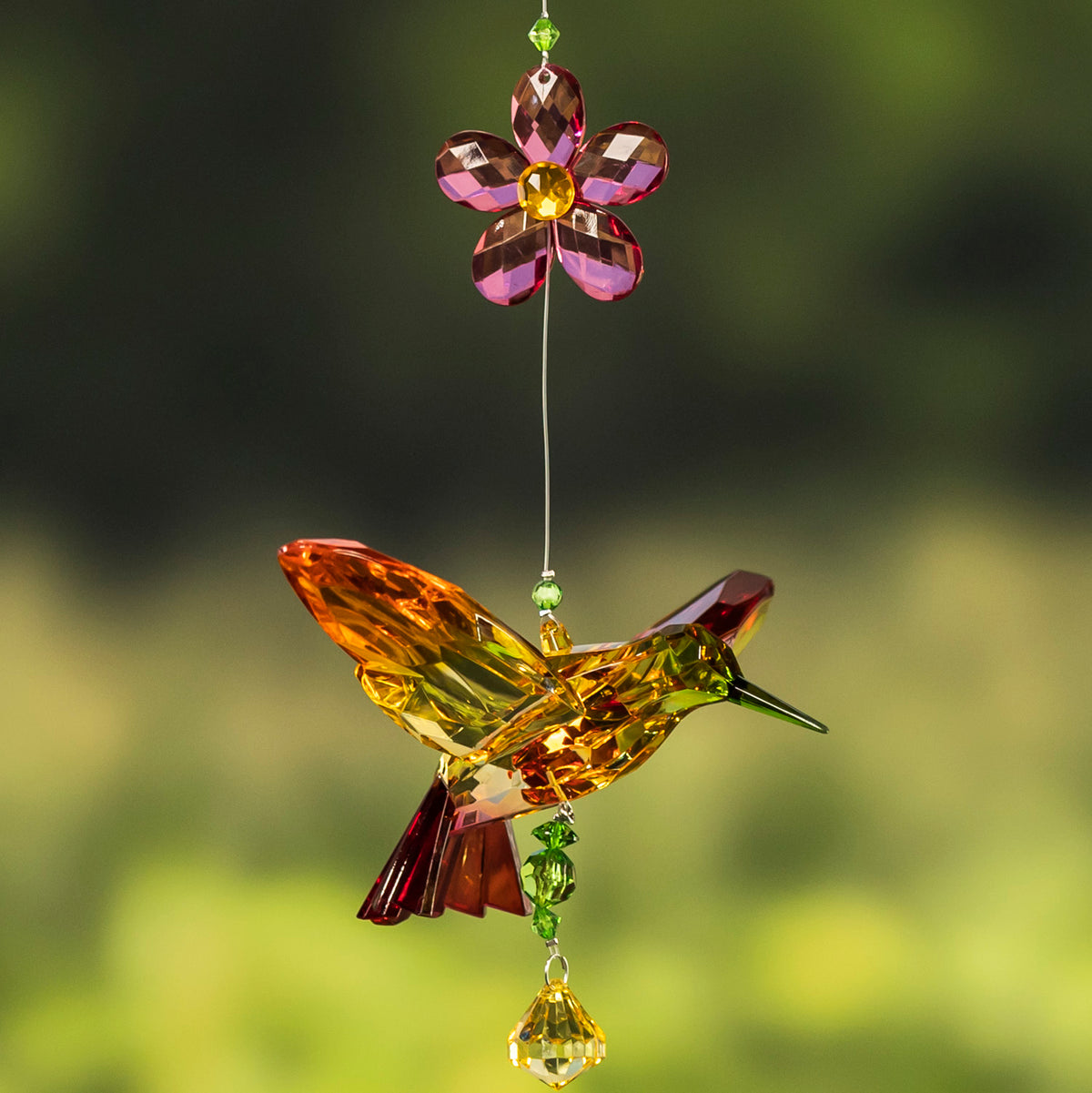 Amber and green faceted hummingbird ornament suspended below a pink flower against a soft green backdrop.