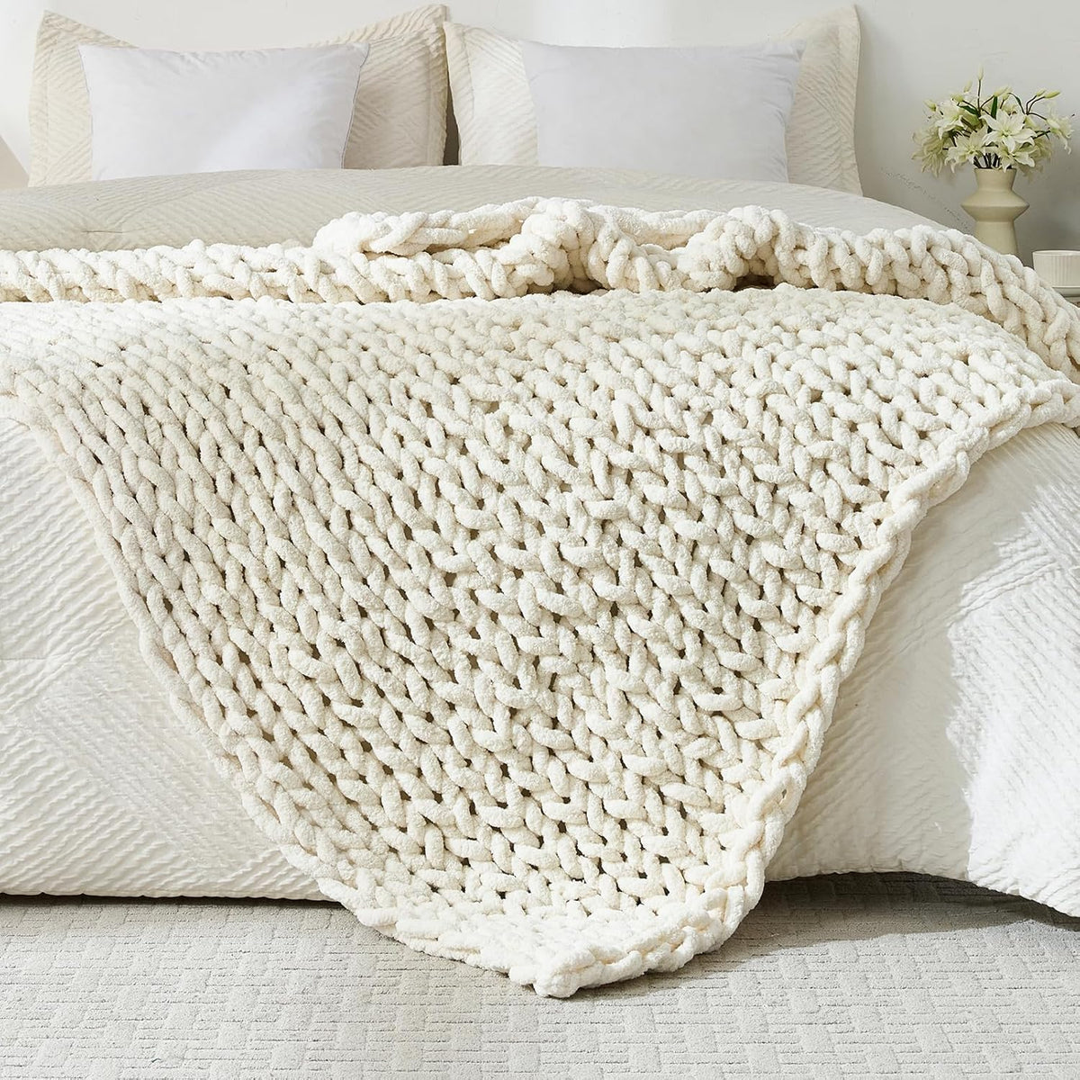 Chunky beige knit throw draped over the foot of a bed with pillows and a vase in background.
