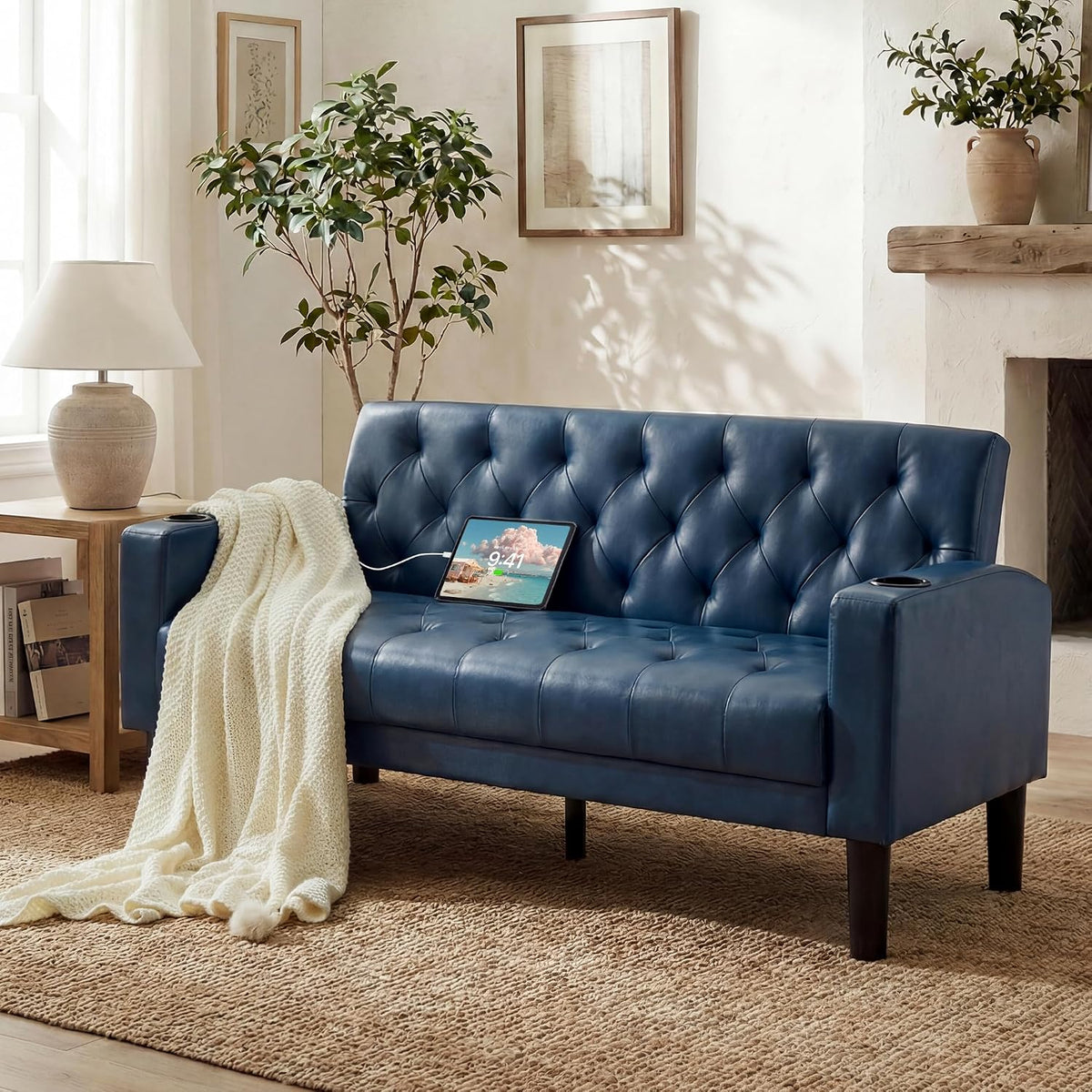 Blue tufted loveseat with USB-charging tablet and cream knit throw draped over the arm in a sunlit room.