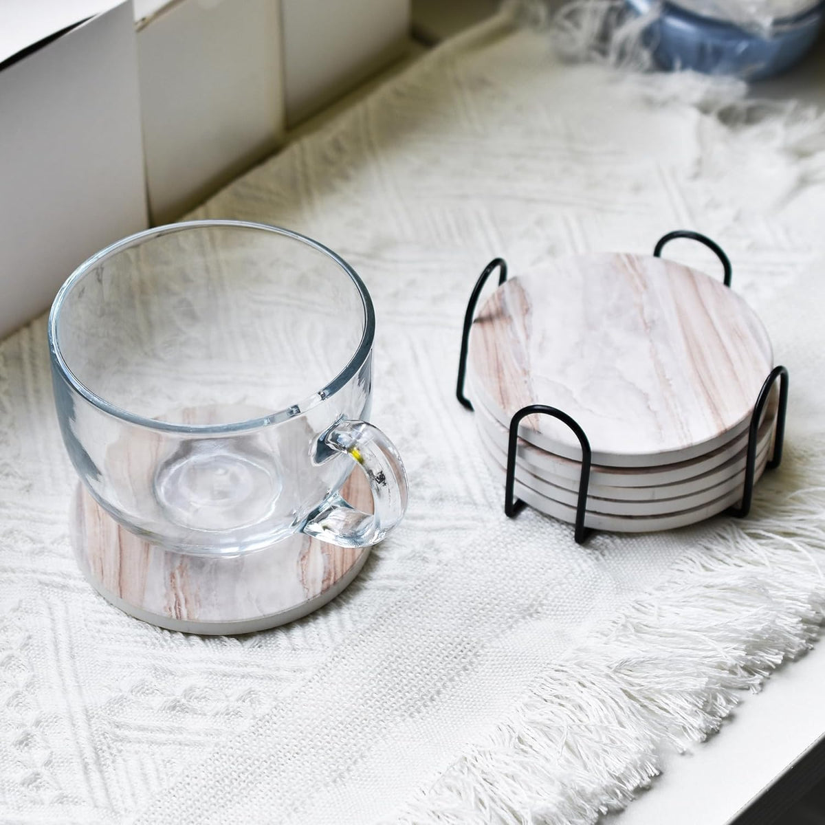 Clear glass mug resting on a marble-pattern coaster next to a stack of coasters in a black metal holder on a textured white r