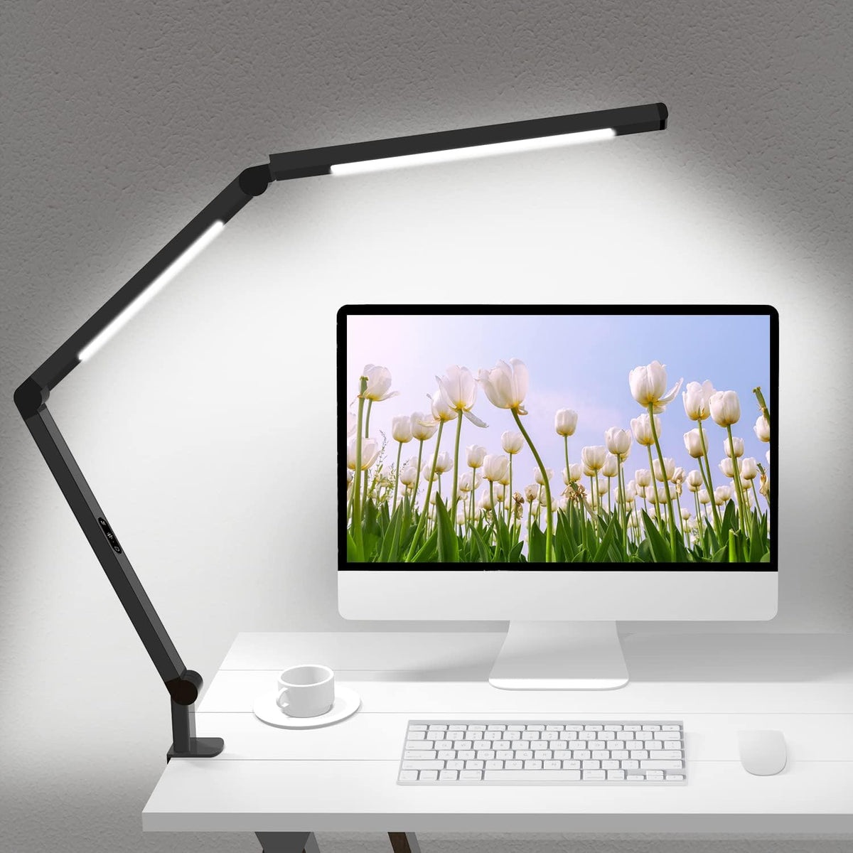 Black dual-light clamp lamp angled over a desktop monitor casting even illumination on the wall behind it