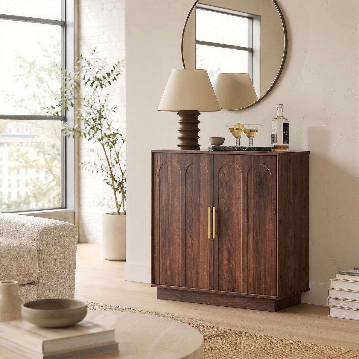 Walnut cabinet topped with a sculpted table lamp and tray of cocktails, reflected in a round wall mirror.