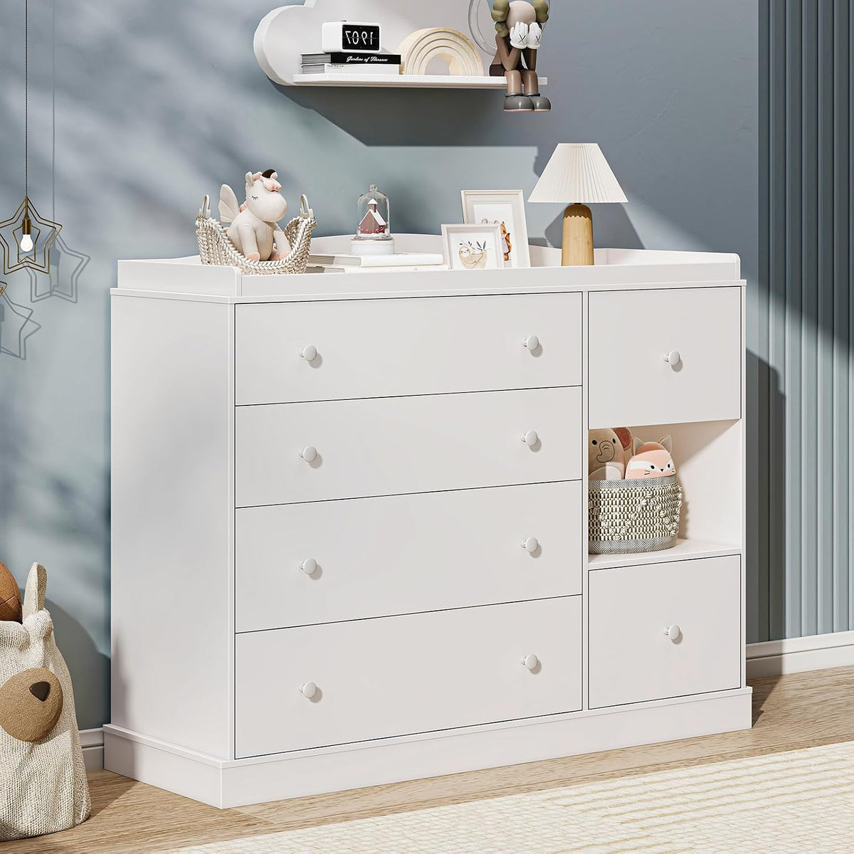 Styled nursery scene showing the white dresser with lamp, basket of toys in the cubby and decorative shelf above.
