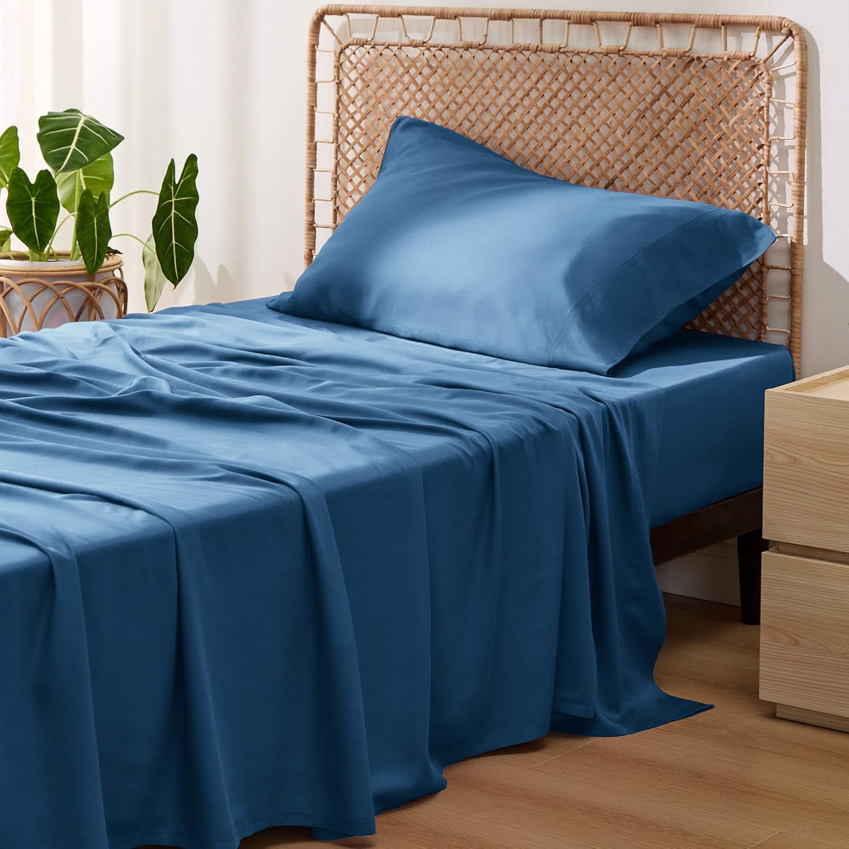 Three-quarter view of a bed with medium blue sheets draped to the floor, a pillow propped against a woven headboard and a