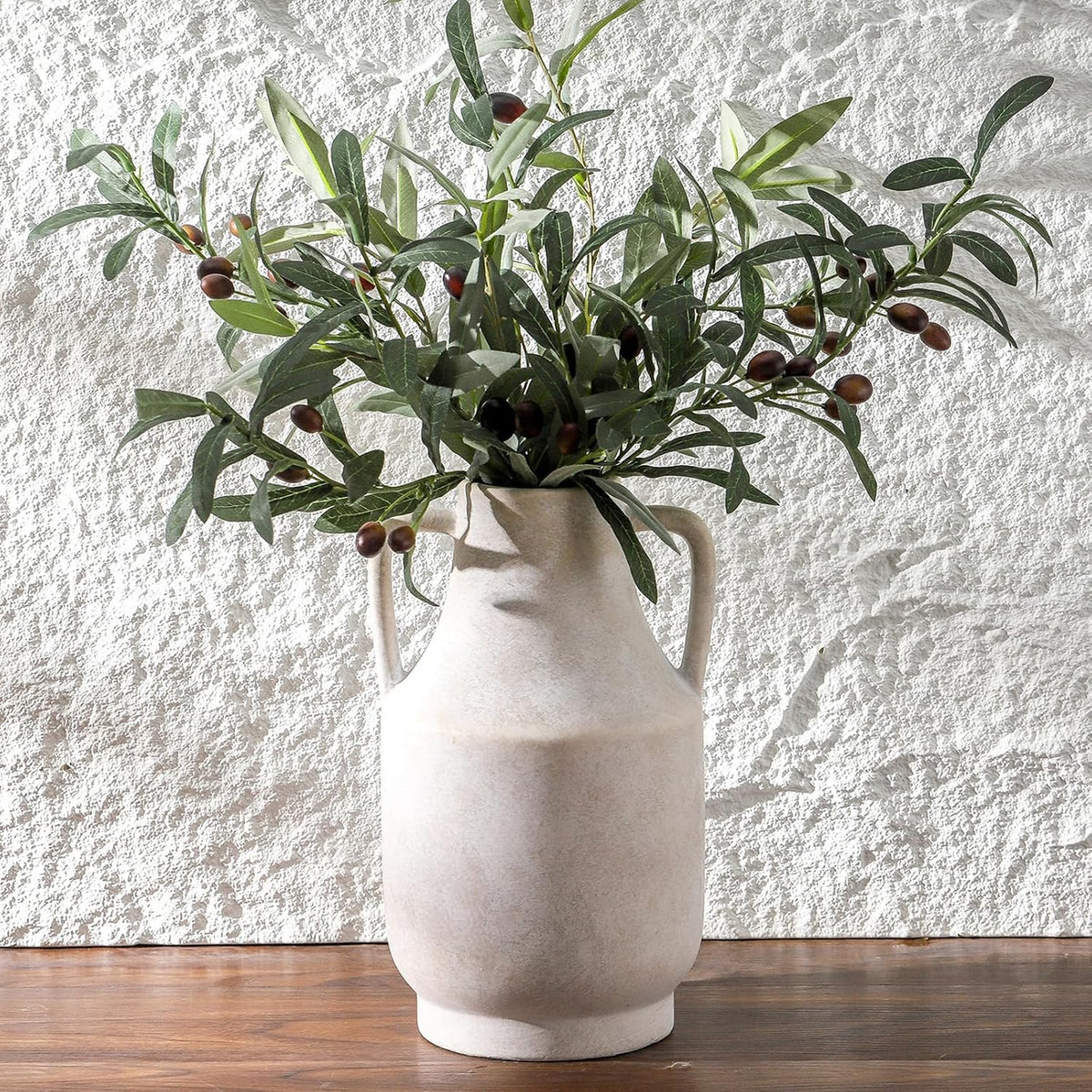 Tall handled beige vase filled with olive branches against a textured white wall