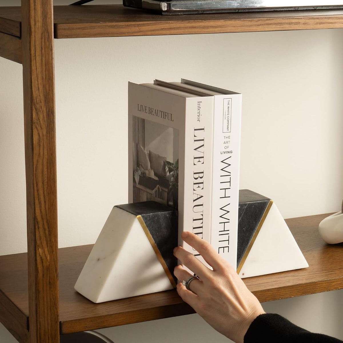 Hand placing white books between gray-and-white triangular marble bookends with gold trim on a wooden shelf.