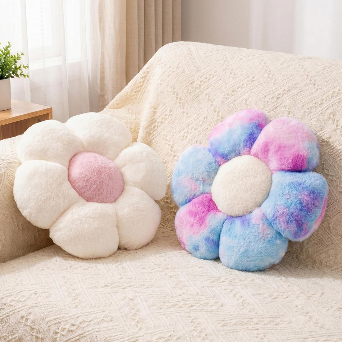 Two plush flower-shaped throw pillows on a cream sofa — one white with pink center, one pastel tie-dye blue/pink with cream