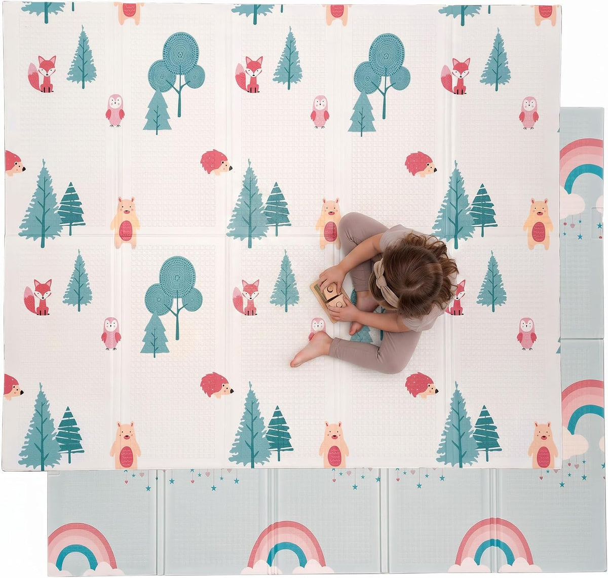 Top-down shot of a child seated on a white play mat showing trees, foxes, owls and bears with a rainbow border visible.